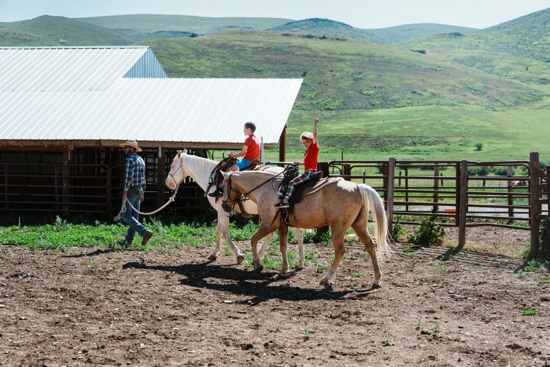 A rancher takes a break from work to lead two horses in the corral as his young daughter and son enjoy a ride.