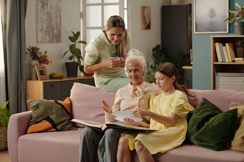 Senior Caucasian woman sitting on sofa with young Caucasian girl looking at photo album, while middle aged Caucasian woman standing behind them smiling and holding smartphone