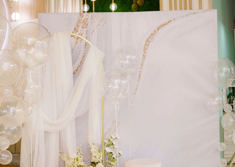 The space features a beautifully arranged backdrop with draped fabric and balloons, perfect for a wedding or party celebration. Fresh flowers add a touch of elegance.
