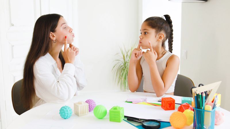 During a therapy session, a speech therapist engages with a child by demonstrating facial movements and articulation techniques in a cheerful environment.