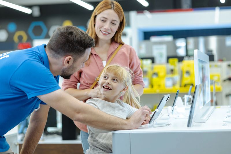 A friendly salesman helps a smiling mother and her daughter explore tablet displays in a bright electronics store. The family chats warmly while interacting with devices, shopping for new tech together.