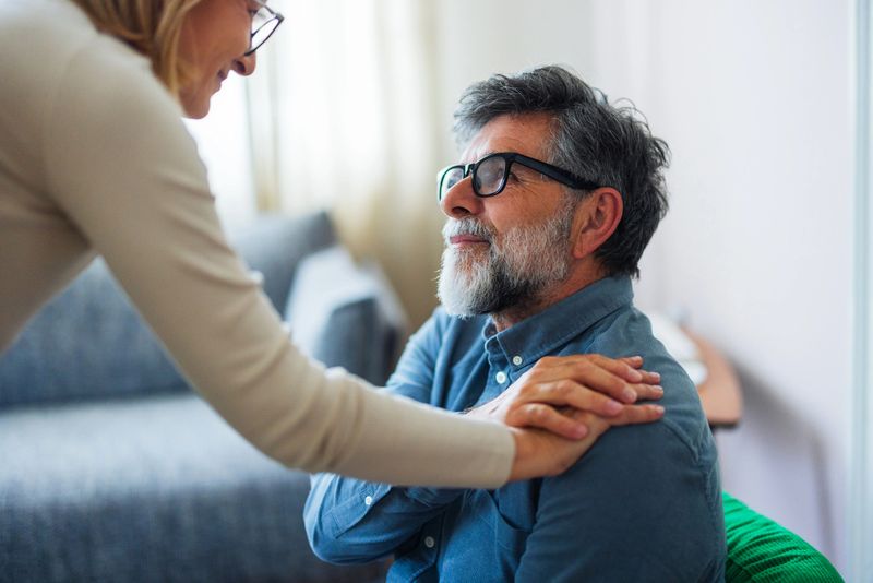 A warm, supportive moment indoors as a woman places her hands on an elderly man's shoulders. He wears glasses and a blue shirt, reflecting trust, care, and connection in a family or caregiver scene.