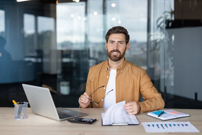 A smiling businessman in a bright office holding glasses with documents and a laptop on the table in front of him.