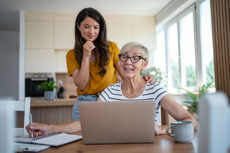 Young adult woman assisting a senior adult mother with online learning and technology while sitting at a kitchen table, fostering an intergenerational bond