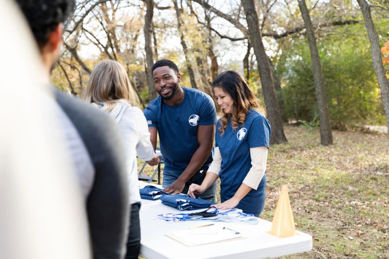 Smiling volunteers hand out items outdoors at a table, demonstrating community engagement and positive teamwork.