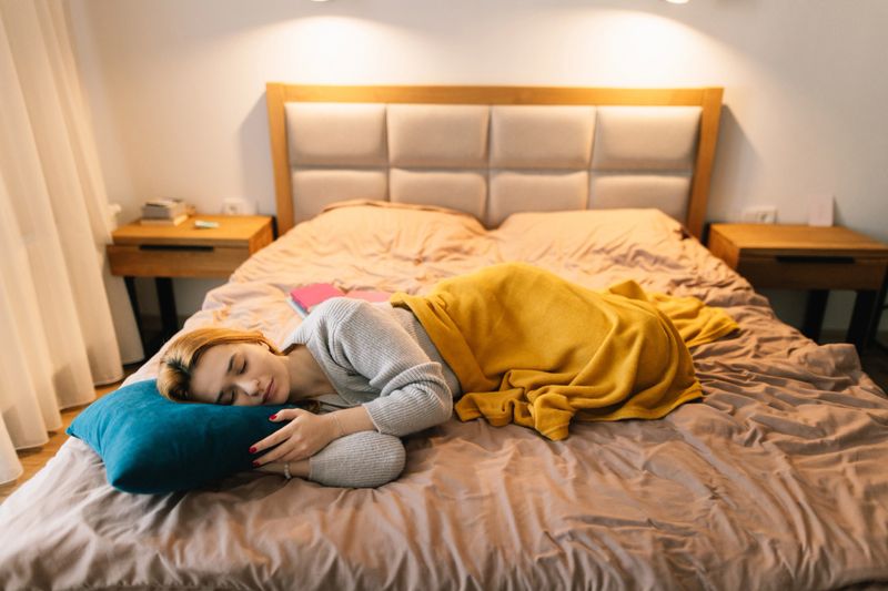 A calm, restful scene of a woman sleeping on a bed with a mustard blanket in a warm, cozy hotel bedroom