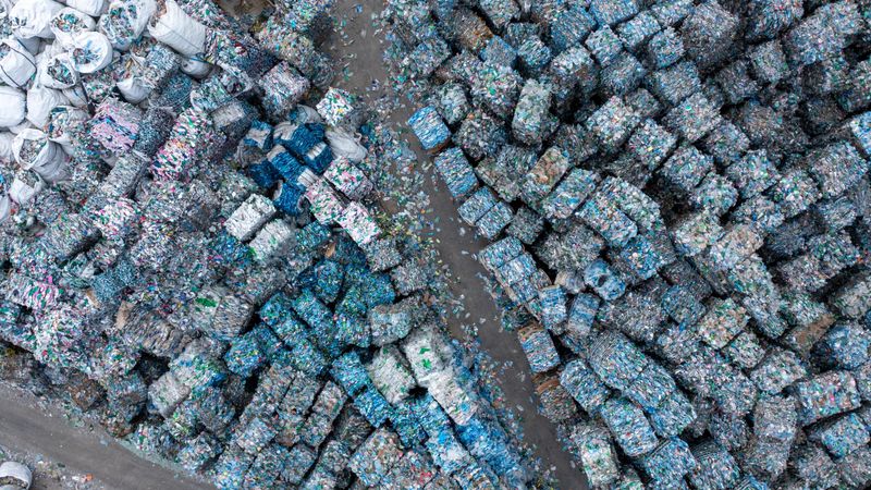 Aerial view of an outdoor recycling waste facility with numerous compressed plastic bales arranged in rows. The site shows organized stacks of colorful plastic waste materials, typical of industrial recycling operations under natural daylight