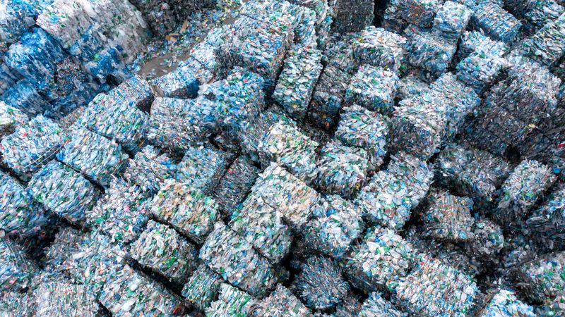 Large compressed bales of mixed recyclable waste stacked outdoors in a waste recycling facility with natural lighting, showcasing industrial recycling processes and environmental waste management