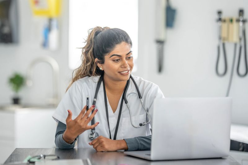 A friendly South Asian nurse sits at a clinic desk, wearing scrubs and a stethoscope, gesturing while talking on a video or virtual meeting beside a laptop, conveying professional care and telehealth readiness.