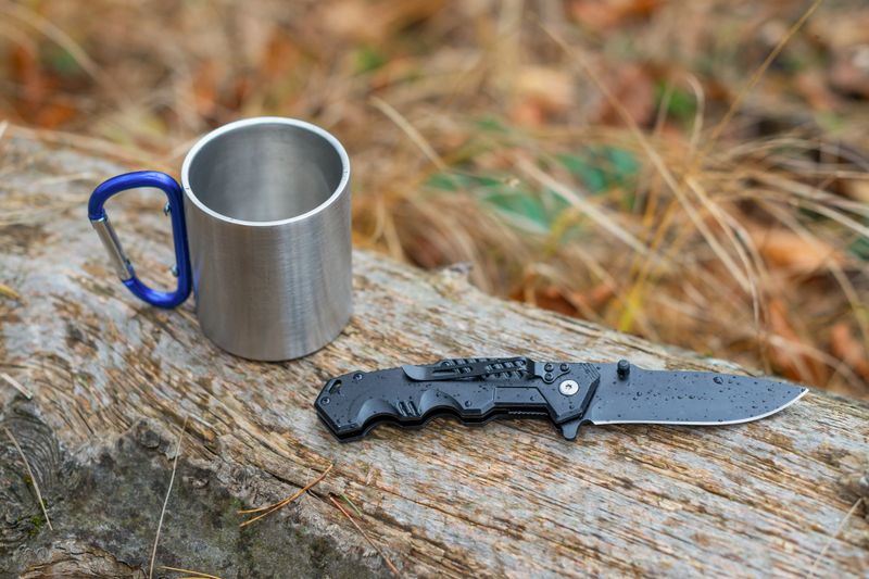 Outdoor camping gear on a rainy day with a folding knife and a metal mug on a wooden log.