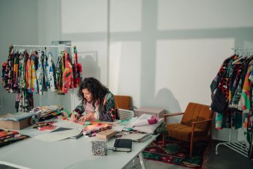 A woman works at a table surrounded by colorful clothing racks.