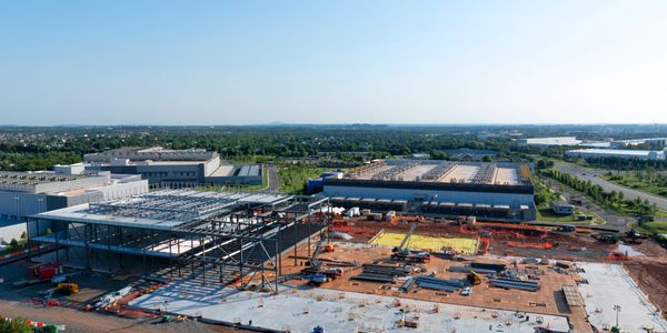 A large construction site with steel framework and nearby industrial buildings.