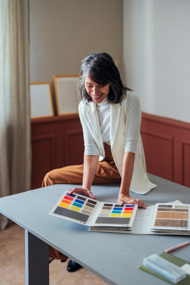A cheerful woman in a white blazer explores color swatches and fabric samples laid out on a gray table, capturing creativity, design thinking, and a stylish home office vibe.