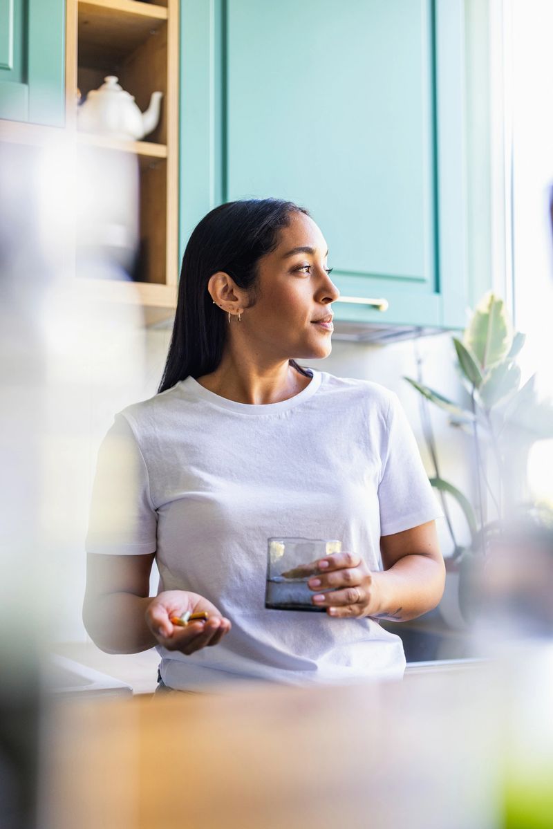 Confident woman stands in a bright modern kitchen with teal cabinets, holding a glass of water and pills. A relaxed, everyday wellness moment captured with natural light.