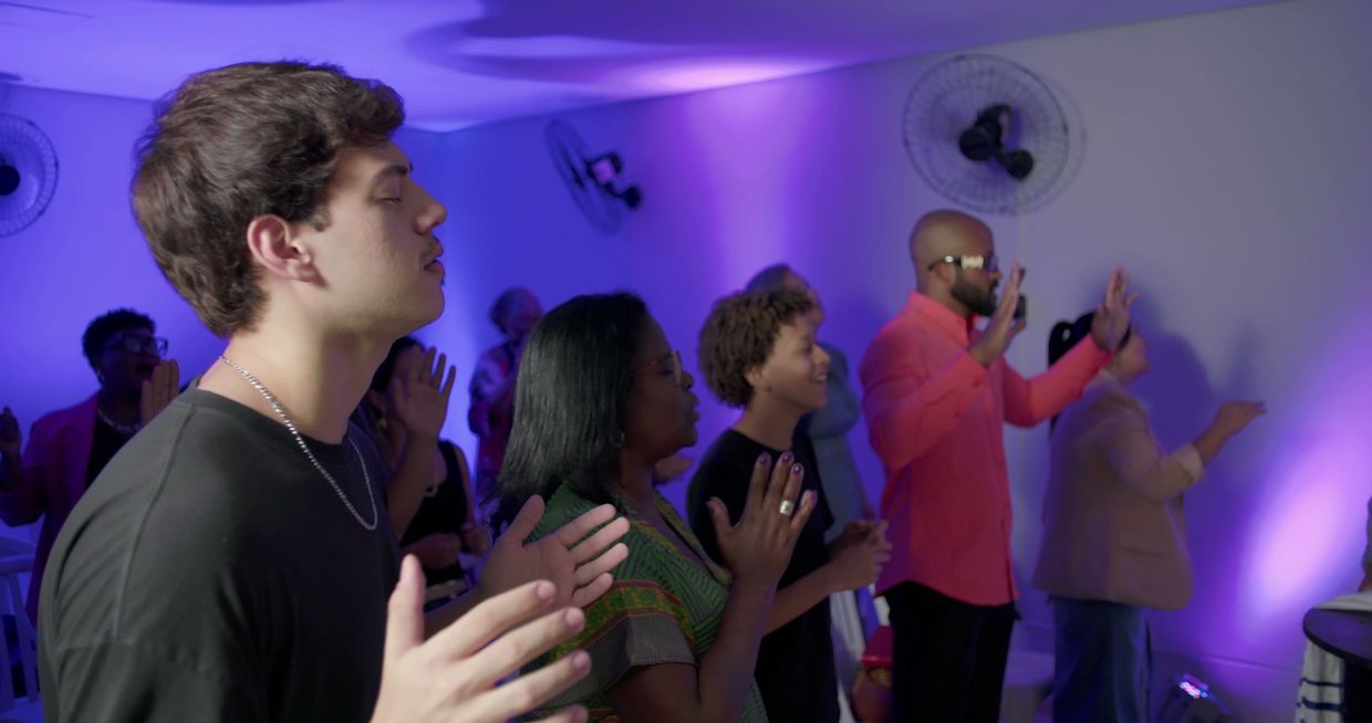 People with closed eyes raising hands in a serene indoor gathering with purple lighting.