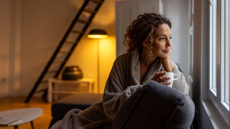 A woman in a soft robe sits by a sunlit window, holding a mug and gazing outside. Warm interior lighting and a cozy home setting convey relaxation and peaceful morning vibes.