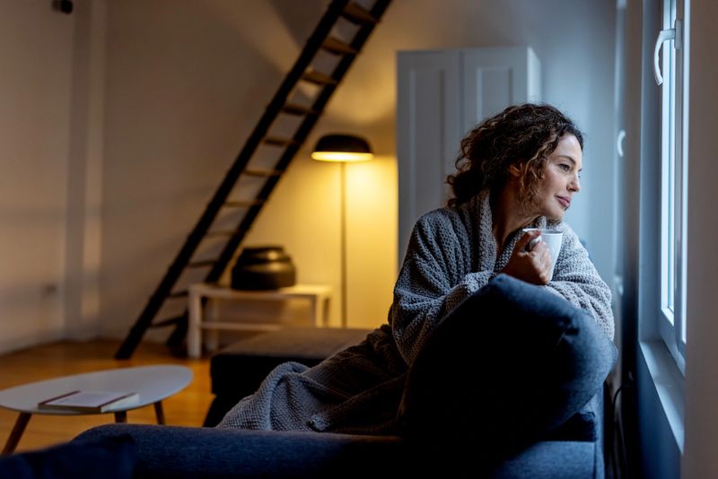 A woman in a plush robe sits on a couch by a sunlit window, holding a mug and gazing outside. Warm interior lighting creates a calm, intimate moment of relaxation.