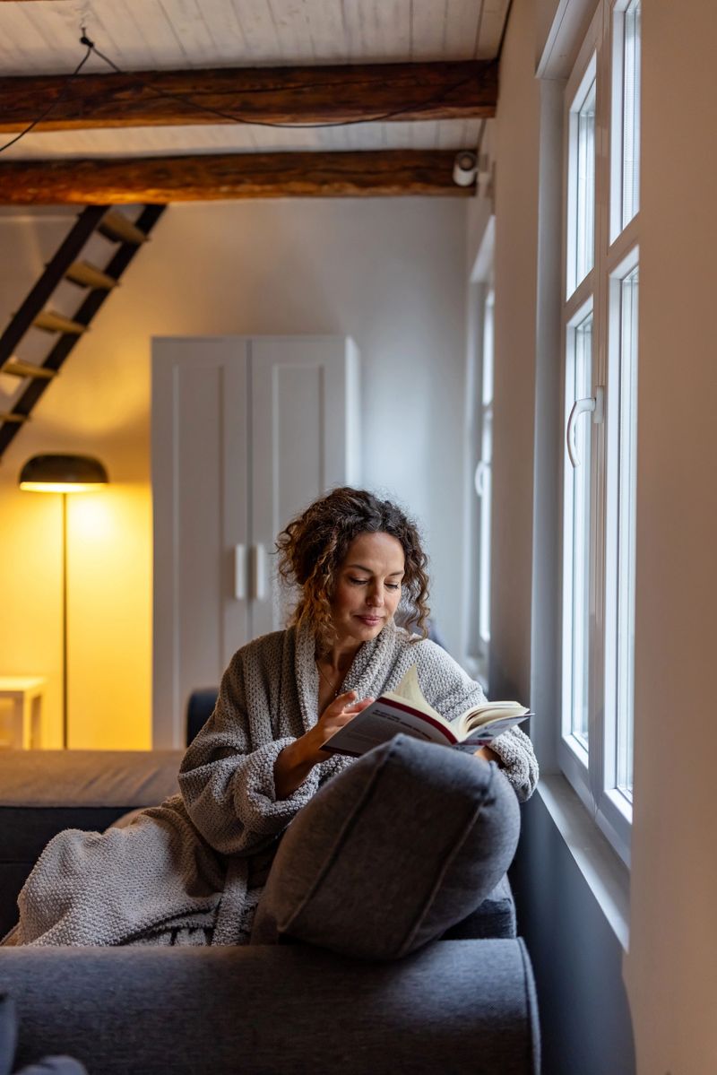 A woman in a textured robe sits on a sofa by a bright window, reading a book in a warm, cozy living room. Natural light and soft interior textures create a calm, intimate mood.