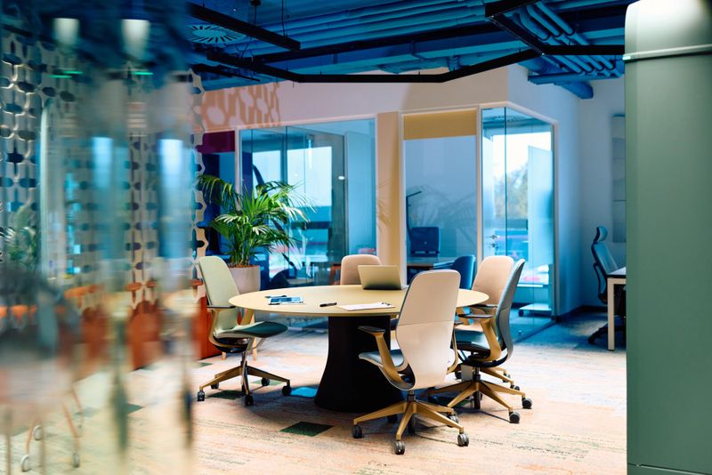 Contemporary open-plan office features a central oval meeting table with a laptop and papers, surrounded by ergonomic chairs and a potted plant, creating a collaborative workspace