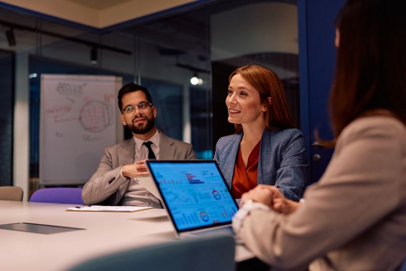 Three adults, a diverse business team, collaborating and discussing financial data on a laptop in a modern corporate board room, working late for a presentation