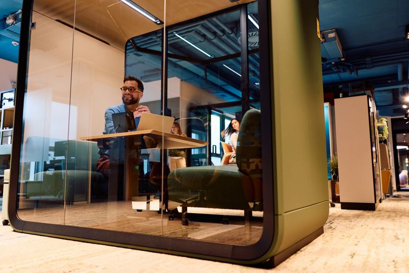 Mid adult businessman smiling at his laptop while seated in a soundproof privacy pod in a modern flexible office space, focused on work and productivity
