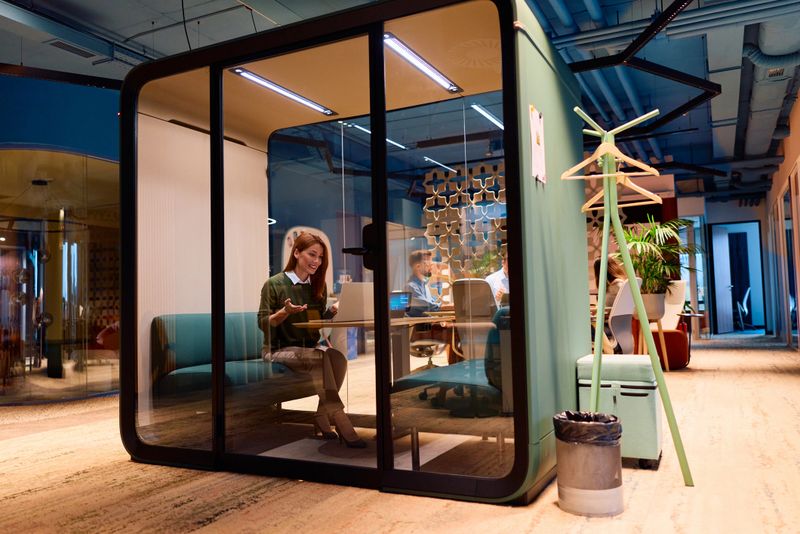 Young professional woman in a soundproof office pod giving a focused laptop video call in a modern coworking space, enjoying privacy and concentrated remote work environment