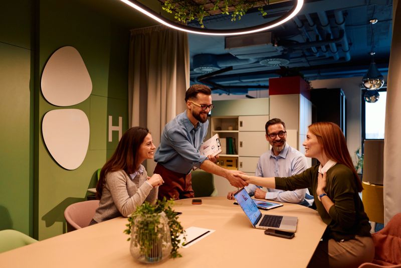 Business colleagues shaking hands across a meeting table in a modern office, celebrating a successful deal or new partnership while conversing with team members