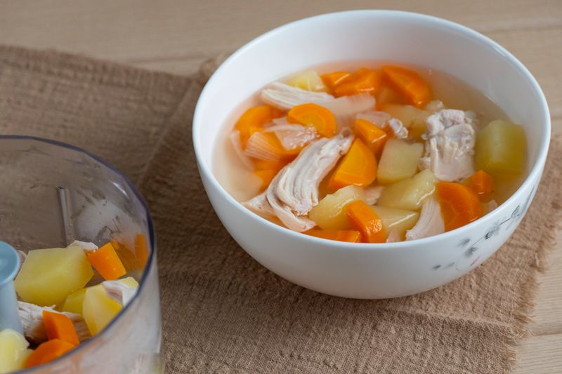 Pieces of boiled chicken, potatoes, and carrots in a bowl and blender cup. Homemade chicken soup preparation on beige textile background