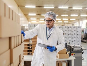 Worker inspecting stacked boxes in a factory with tablet in hand.
