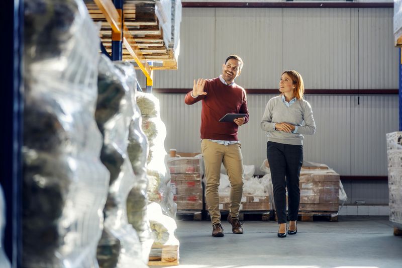 Two business partners walking through a large warehouse, a man holding a digital tablet and talking with a woman