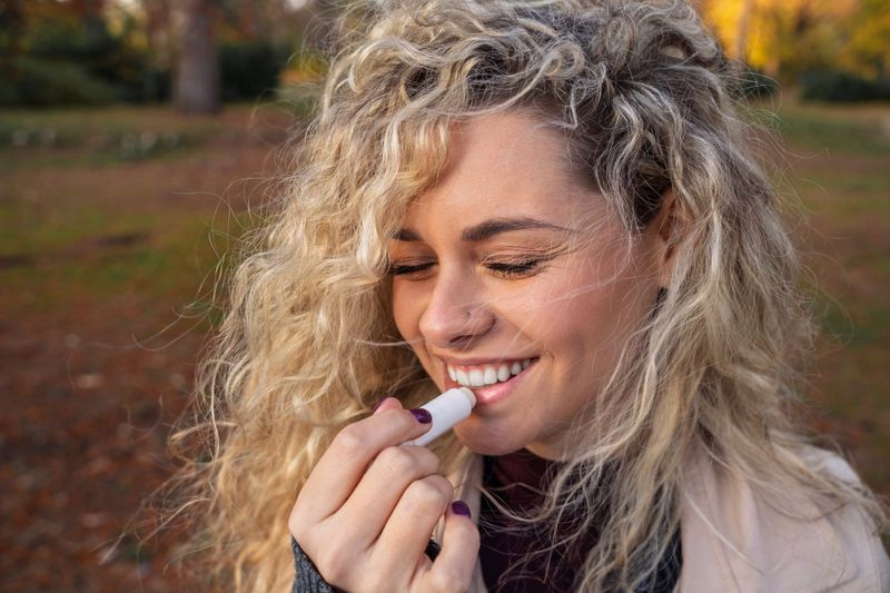 Gorgeous natural young girl with beautiful curly hair putting on lip balm on her mouth while smiling , outdoors in the park