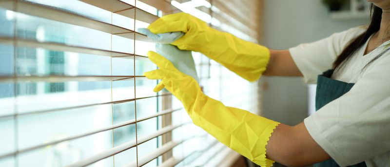 A hand wearing a protective yellow glove carefully wipes wooden Venetian blinds with a microfiber cloth illuminated by soft natural light indoors.