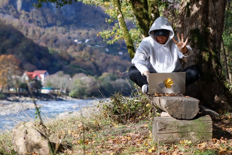 Person in hoodie showing peace sign with laptop in autumn landscape. Hooded individual seated under tree makes victory gesture, silver laptop on log with view of flowing river and colorful foliage.