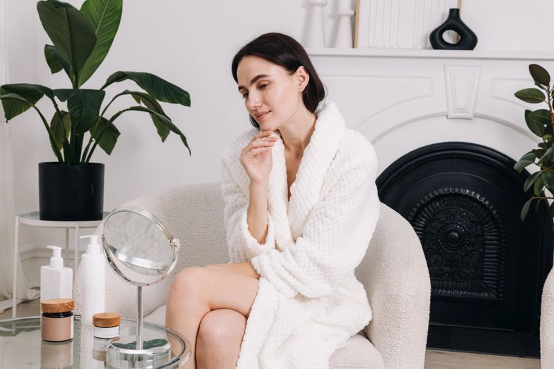 Young woman in a terry robe thoughtfully applies her skin care products, use face cream while seated by a mirror, enjoying her morning pampering routine in bathroom or living room