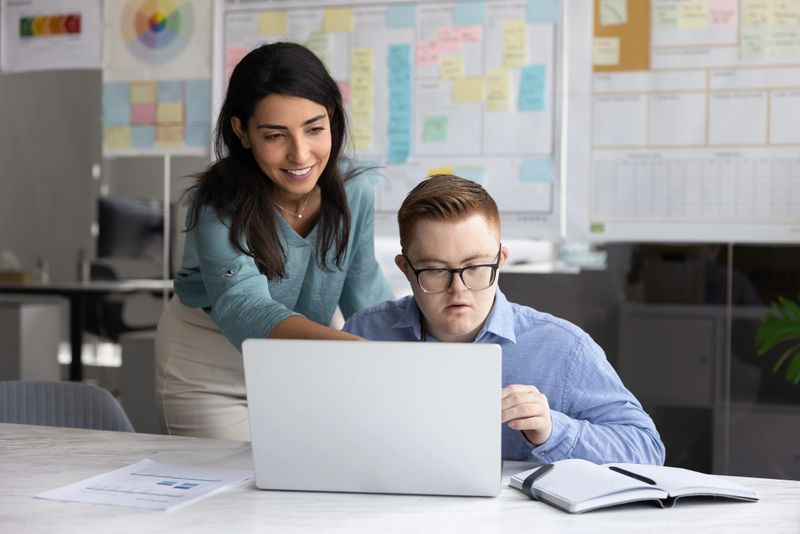 Friendly young Latin mentor woman training male intern with Down syndrome, explaining task, showing online content on laptop, pointing at screen, explaining work application