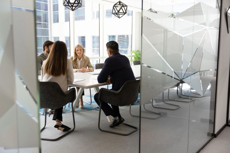 Through doorway view at business team talking at large meeting table in office collaboration room behind glass wall, discussing teamwork strategy, partnership, agreement. Wide candid shot