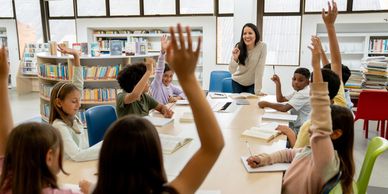 Children raising hands in classroom with teacher.