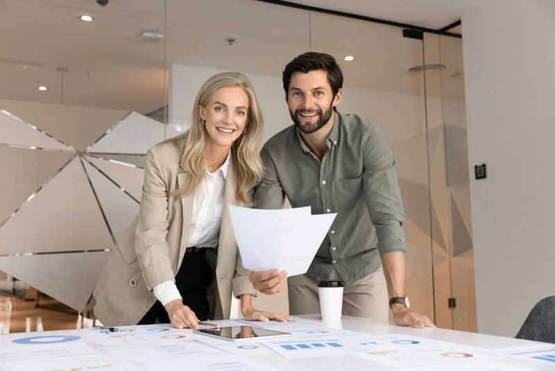Couple of happy marketing professionals working together, posing at table with heal of paper statistic reports, analyzing company sales, project data, looking at camera, smiling