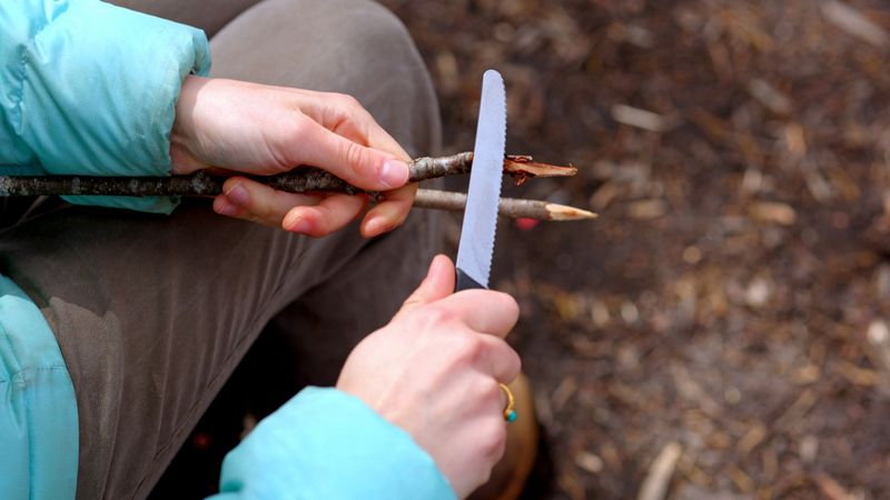 Detail of young woman cutting stick with knife at campsite