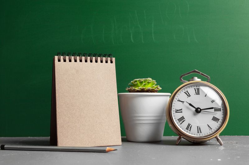 Notepad and clock sit beside a potted plant on a desk with a green chalkboard in the background.