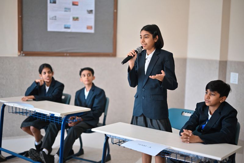 Students in formal uniforms sit in teams while one participant stands with a microphone to present her arguments, as the opposing team listens and prepares their rebuttal.
