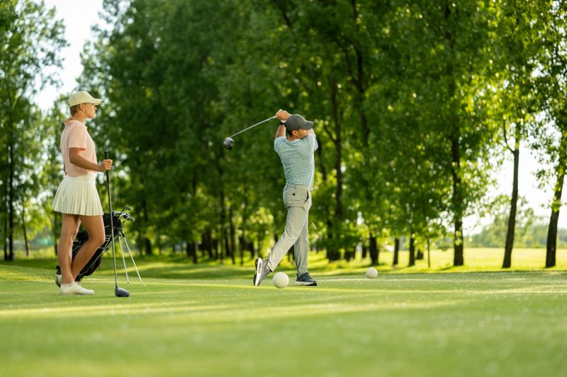 Father swinging a golf club on a sunny tee box while daughter watches attentively. They are surrounded by lush green trees, creating a vibrant outdoor setting. Golf clubs are placed nearby, indicating an active day of golfing.