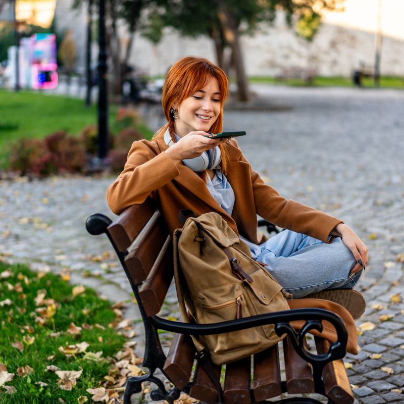 Young woman smiling, holding smartphone near mouth, sending voice message while sitting outdoors on a park bench during autumn