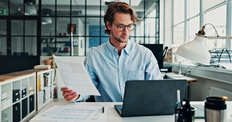 Accounting, documents and laptop with business man at desk in office for budget or finance. Computer, paperwork and reading with employee in financial workplace for bookkeeping or wealth management