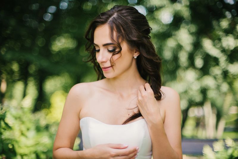 A radiant bride with elegant makeup and curly brown hair smiles gently in a lush park her elegant makeup and wavy brunette hair, natural light highlights her features against a beautiful green bokeh background. Wedding luxury portrait