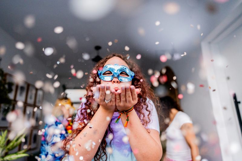 A vibrant portrait of a teenage girl celebrating Carnival, wearing a decorative blue mask and blowing confetti with excitement. The image captures the playful spirit of the festival, with colorful paper pieces floating around her in a dynamic, joyful scene.