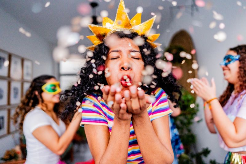 A vibrant and energetic scene capturing the essence of Carnival celebration, with a young woman in a colorful striped shirt and golden crown blowing confetti. Her friends can be seen in the background, creating a festive and playful atmosphere filled with joy and excitement.
