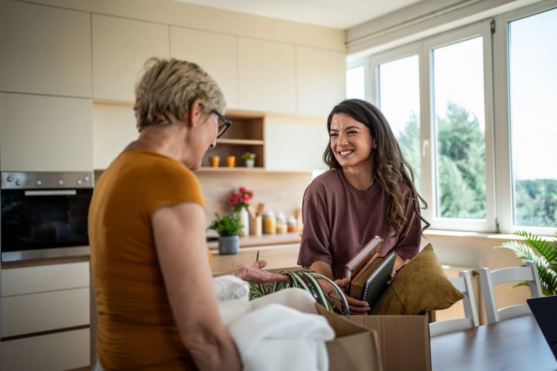 Mother and daughter smiling, talking, and unpacking boxes, helping each other move into a new home kitchen, sharing a moment of togetherness and new beginnings