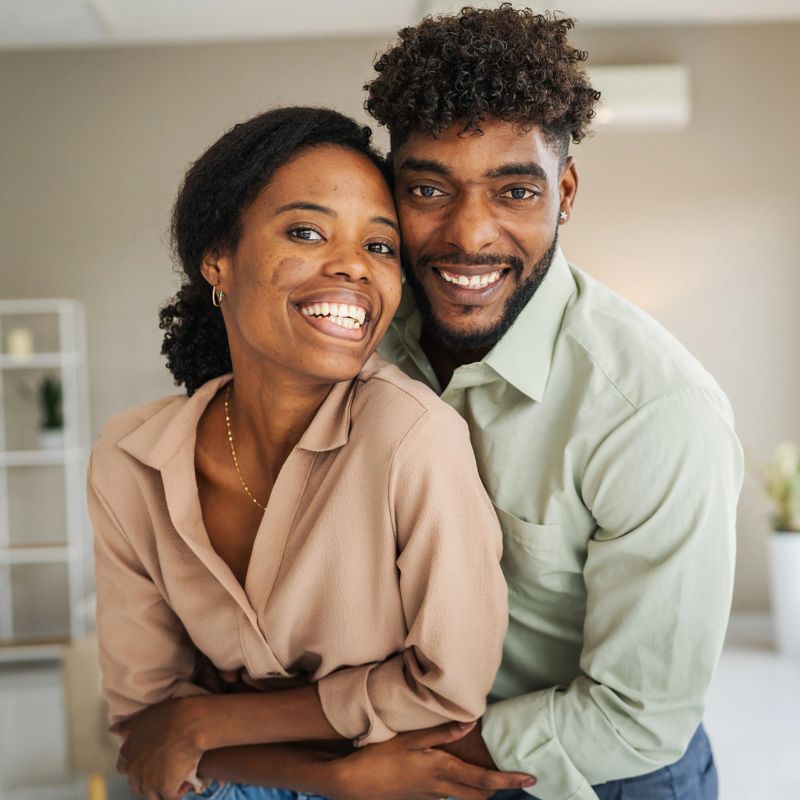 Happy young black couple embracing and smiling at the camera in a bright modern apartment, sharing affection, warmth and joyful connection in their cozy living space