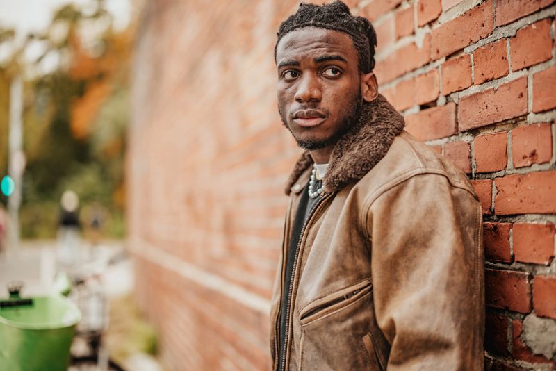 A young person stands against a brick wall in Berlin, taking in the vibrant atmosphere. Surrounded by greenery and city life, they embrace a moment of joy and contemplation outdoors.